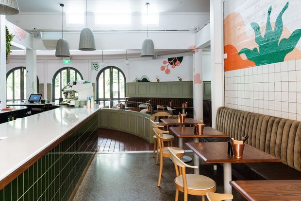 A bright, eclectic contemporary café interior with a long green-tiled service counter, polished light benchtop, arched windows, hanging woven pendant lights, and rows of timber tables and chairs. A ribbed upholstered banquette runs along a tiled feature wall with colourful abstract artwork, while pale green cabinetry and dark timber finishes add contrast.