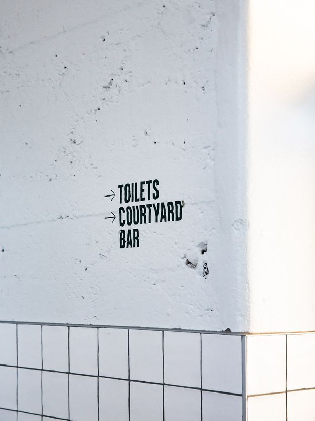 Close-up of a white textured plaster wall with black directional signage reading “TOILETS,” “COURTYARD,” and “BAR.” The lower portion is finished with a grid of white square tiles and dark grout lines, creating a simple commercial wayfinding detail.