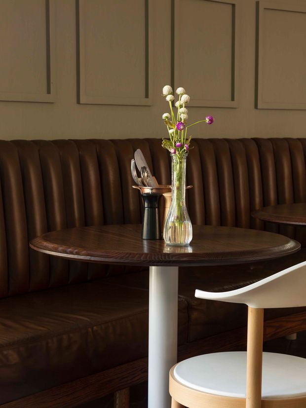 A warmly lit restaurant or café booth with a dark brown vertically channelled leather banquette, a round dark timber table on a white pedestal base, and a white-and-light-wood chair in the foreground. Decorative framed wall panels and a small vase of flowers create a refined, intimate dining setting.