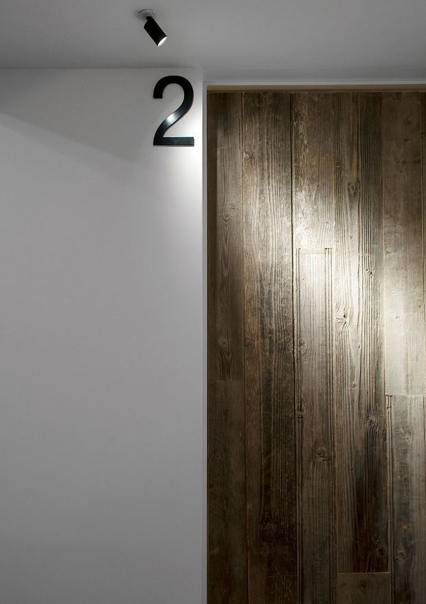 A minimalist interior corridor or entry area featuring a tall rustic timber-clad door or wall panel, a large black number 2 mounted on a white wall, and a small black directional spotlight. The contrast between smooth white plaster and weathered vertical wood creates a simple, contemporary-industrial look.