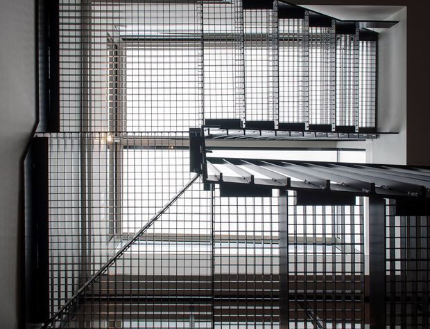 Looking upward through a multi-level interior stairwell or atrium, with steel stair flights, dark metal railings, and extensive square-grid metal screens or balustrades. Bright daylight filters through the tall glazed or screened void, creating a dramatic geometric composition.