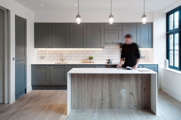 A bright contemporary kitchen with a large central island, dark grey shaker-style cabinetry, white subway-tile splashback, pale timber-look flooring, and a timber-faced island. Three pendant lights hang above the island, with large black-framed windows providing natural light.