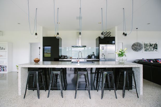 Bright open-plan kitchen dominated by a large speckled stone island with seating for several people. The room features black flat-panel cabinetry, integrated appliances, a pale tiled or glass splashback, stainless-steel tapware, polished concrete or terrazzo-style flooring, pendant lights with exposed cords, and a visible living area beyond the island.
