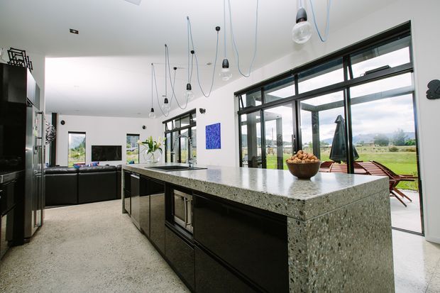 Open-plan contemporary kitchen and living area with a large dark island featuring a terrazzo-style benchtop, extensive black-framed glazing, hanging pendant lights, and views to a landscaped outdoor area. A black sectional sofa and television occupy the adjoining living zone.