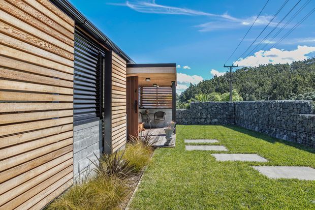 Exterior side courtyard of a contemporary single-storey building, featuring horizontal timber cladding, dark louvred screening, concrete block detailing, and a small covered outdoor seating area. A lawn with rectangular concrete stepping slabs leads alongside a gabion stone boundary wall, with dense vegetation beyond.