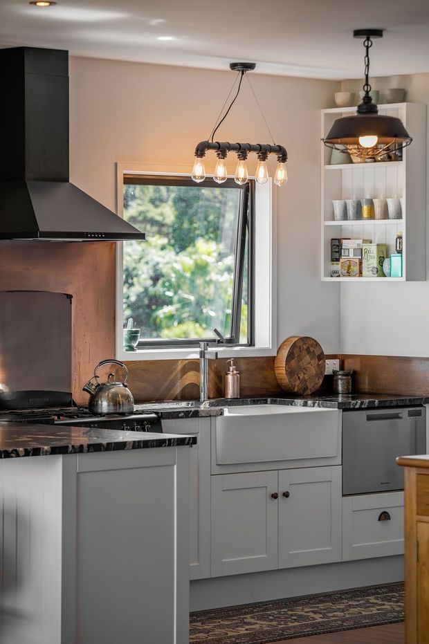 A warm, traditional-style kitchen with pale grey shaker cabinetry, dark polished stone benchtops, a timber-toned splashback, black rangehood, stainless-steel appliances, open shelving, and industrial-inspired pendant lighting. A window brings in greenery and natural light, while a patterned runner adds warmth to the space.