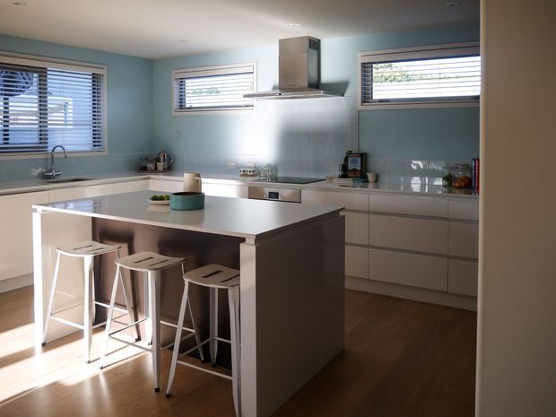 A bright, contemporary kitchen with pale blue walls, white cabinetry, a large white island with integrated seating, stainless-steel rangehood and appliances, and timber flooring. Horizontal windows with white blinds bring in strong natural light.