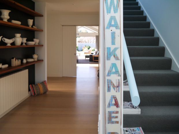 A bright residential hallway with warm timber flooring, open dark timber display shelves filled with ceramics, and a white radiator beneath. A carpeted staircase with a pale blue handrail rises on the right, while a glazed doorway at the end leads toward a sunlit living area. A tall decorative stair panel displays colourful lettering.