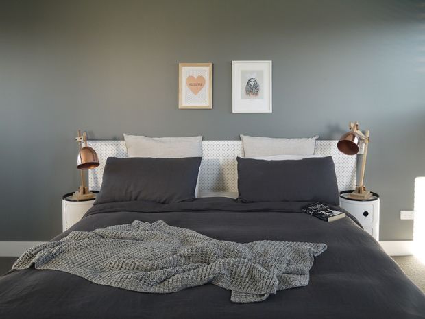 A stylish bedroom with a large dark upholstered or fabric-covered bed dressed in charcoal bedding, oversized dark pillows, and a chunky grey knitted throw. The bed is positioned against a deep grey feature wall with patterned white pillows, matching white bedside tables, and brass-and-brown adjustable lamps. Two framed artworks hang above the bed, and daylight enters from the right.