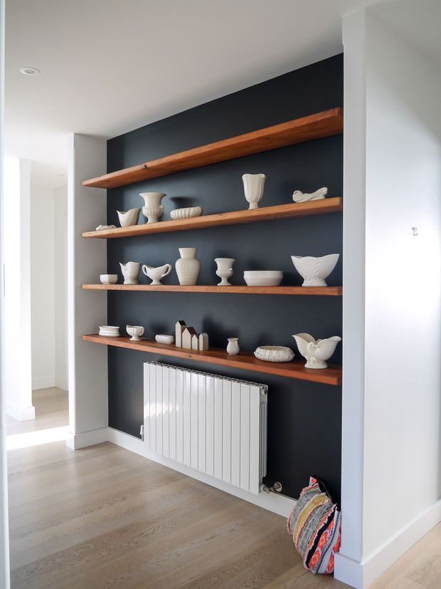 Interior view of a bright hallway or living-area niche featuring a dark charcoal feature wall with four long floating timber shelves displaying white ceramic vessels and decorative objects. A white vertical-panel radiator sits below the shelving, with pale timber flooring, white walls, and open circulation space leading to another room.