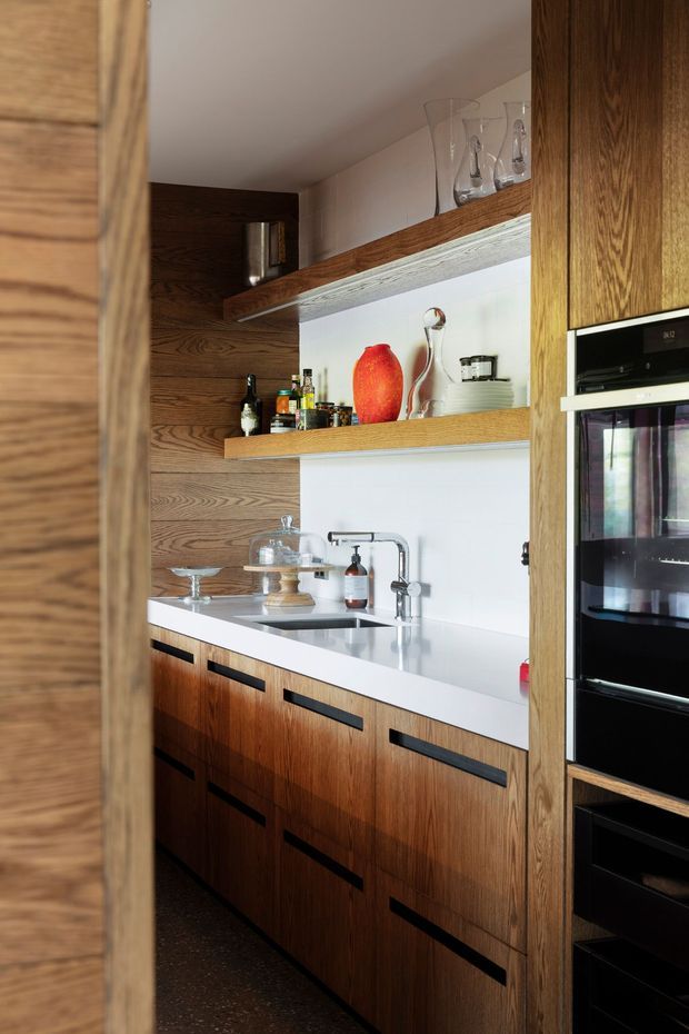 A compact, richly detailed kitchen or butler's pantry with dark timber wall panelling, matching wood cabinetry and open display shelving. The space features a white benchtop and white backsplash, undermount sink with chrome tapware, integrated black appliances, and long black recessed drawer pulls. Glassware and decorative objects are displayed on the shelves, creating a warm, refined mid-century-inspired interior.