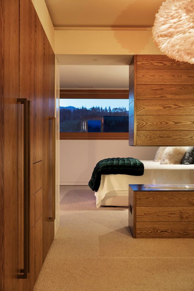 Warmly lit bedroom viewed through a timber-lined entry or wardrobe zone. The room features extensive dark timber built-in cabinetry, a low bed with cream bedding and dark green throw, beige carpet, a timber-framed window overlooking trees and hills, and a large textured pendant light. The design is refined and contemporary with strong natural wood detailing.