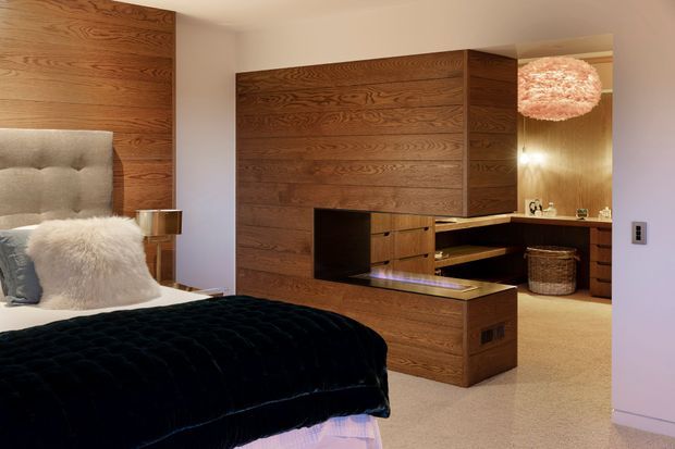 A warm, sophisticated bedroom with a large bed in the foreground, upholstered tufted headboard, white faux-fur throw, and dark navy or black quilt. A substantial dark timber feature wall and built-in joinery conceal and frame an open dressing or storage area, with drawers, shelving and a low partition. The room has soft beige carpet, white walls, warm timber surfaces, brass-toned lighting, and a textured spherical pendant visible near the adjoining dressing area.