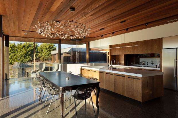 A refined open-plan kitchen and dining area with extensive floor-to-ceiling glazing, a dark dining table, woven dining chairs, and a large sculptural branch-like pendant light. The kitchen features warm timber cabinetry, a long island with a pale stone benchtop, integrated appliances, and a richly lined timber ceiling with exposed beams. The space opens visually to a landscaped exterior and receives abundant natural light.
