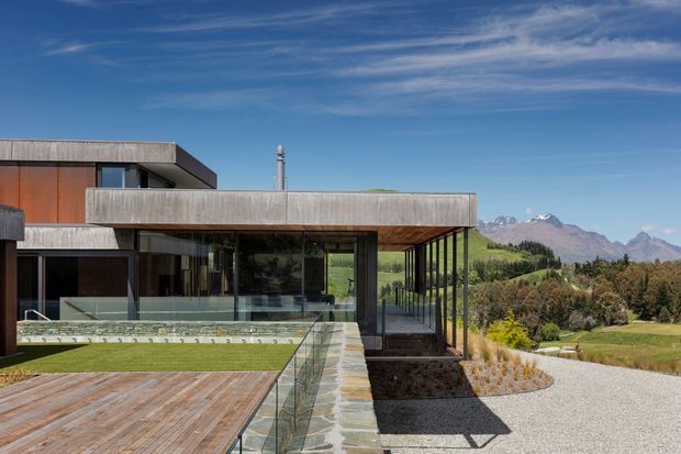 A contemporary flat-roofed house set in a scenic mountainous landscape. The building features extensive floor-to-ceiling glazing, strong horizontal concrete forms, dark window frames, warm timber cladding, a timber deck, glass balustrades, and a stone-paved walkway. A landscaped lawn and gravel forecourt frame the residence, with rugged snow-dusted mountains visible beyond.