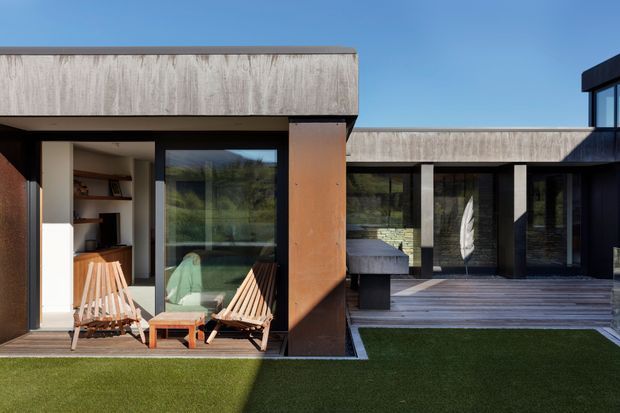 A modern single-storey courtyard residence viewed from the outdoor lawn. The house has flat roof volumes, dark-framed floor-to-ceiling glazing, weathered concrete or cementitious cladding, and rust-toned corten-like metal panels. A timber deck connects the glazed living areas, with lounge chairs and a built-in outdoor feature visible in the courtyard.