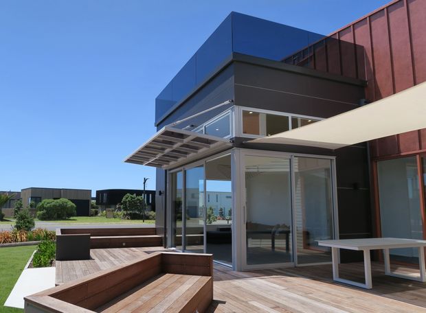 Contemporary multi-level building exterior with a dark charcoal and blue-clad projecting volume, extensive glazed corner doors and windows, a slatted metal awning, and a large white shade sail. The image focuses on a timber deck terrace with built-in timber seating, an outdoor dining table, landscaped garden edges, and a glimpse of interior furnishings through the glazing. Clear blue sky and neighbouring modern buildings form the background.