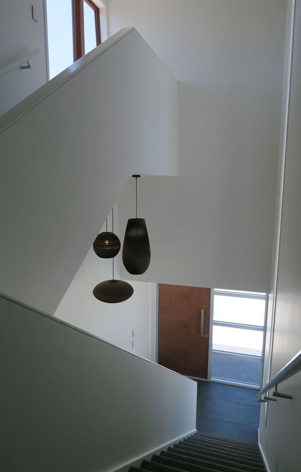 A tall, minimalist residential stairwell viewed from the upper stairs, with white walls and angular stair partitions framing a double-height void. Three dark sculptural pendant lights hang above the stairwell. A dark timber entry door with a long vertical handle and adjacent glazing is visible at the lower landing, with grey tiled flooring and simple metal handrails.