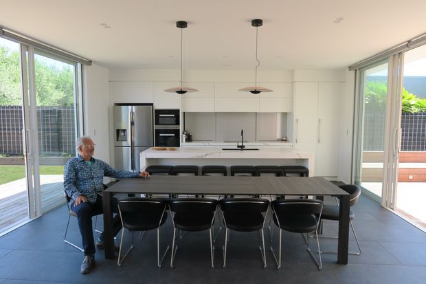 Open-plan contemporary kitchen and dining area with a long dark dining table and black chairs in the foreground. A white minimalist kitchen with integrated cabinetry, stainless-steel refrigerator, built-in ovens, island bench, and pendant lighting sits behind it. Extensive floor-to-ceiling glazed sliding doors open to a bright outdoor terrace and garden, creating an airy indoor-outdoor connection.