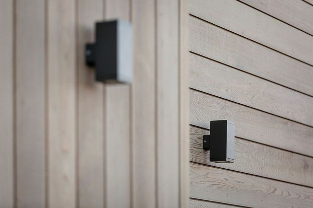 Close-up architectural detail showing two minimalist black rectangular wall-mounted light fixtures against pale timber cladding: vertical board-and-batten-style panels on the left and horizontal weatherboard-style panels on the right.