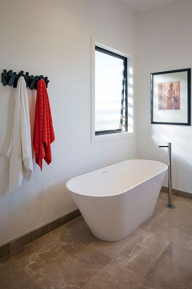 Bright, modern bathroom featuring a freestanding white oval bathtub, wall-mounted towel hooks with white and red towels, a narrow black-framed window, polished chrome floor-mounted bath tap, framed artwork, and large brown-beige stone-look floor tiles.