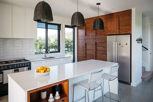 Bright contemporary kitchen with a large white island and integrated seating, white flat-panel cabinetry, stainless-steel appliances, dark woven pendant lights, and a striking full-height timber feature wall with concealed cabinetry. A stairway is visible beyond the kitchen.