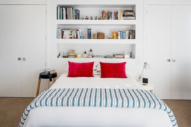 Bright, compact bedroom with a neatly made bed, white built-in wardrobes and recessed open shelving filled with books and decorative objects. The bed is styled with vivid red cushions and a blue-and-white striped throw, while small bedside stools and lamps sit on either side. The space has a clean, contemporary built-in design and a neutral palette with colourful accents.