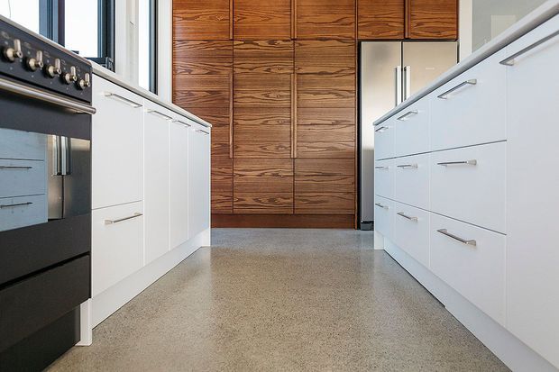 A contemporary galley kitchen viewed from the central aisle, with white flat-panel cabinetry on both sides, integrated appliances, dark timber veneer cabinetry along the rear wall, and a polished speckled concrete or terrazzo-like floor. The room has a clean, functional layout with strong linear geometry and natural light from the side windows.