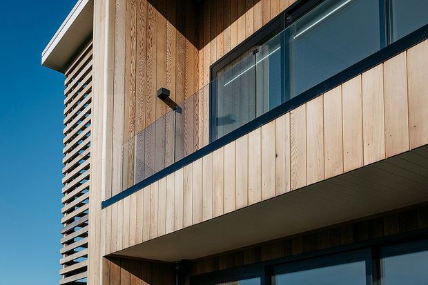 Close-up exterior view of a contemporary multi-storey building clad in pale vertical timber boards, with projecting balconies, clear glass balustrades, dark metal framing, and a prominent louvred screen along the left side. Strong sunlight creates crisp shadows against the timber facade and glazing.