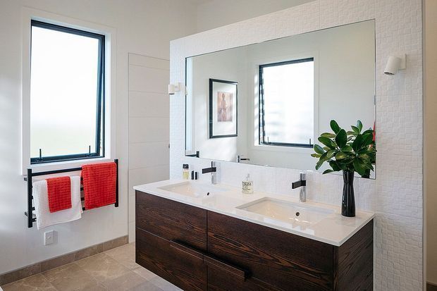 Bright contemporary bathroom with a double floating vanity in dark timber cabinetry, white integrated basins, matte black tapware, and a large frameless mirror. White textured wall tiles, pale floor tiles, black-framed windows, and red towels add contrast, while a potted plant provides a natural accent.