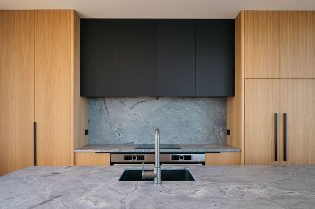 Contemporary kitchen viewed from the island, featuring full-height light timber cabinetry, matte black overhead cabinets, a large grey stone island and matching splashback, integrated cooktop and sink, and minimalist black hardware.