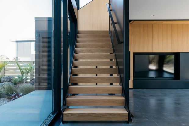 Contemporary interior view of an open-riser timber staircase with black metal framing and handrail, positioned beside floor-to-ceiling glazing. A timber-panelled wall with a built-in black television unit is visible to the right, while landscaped exterior planting is seen through the glass.