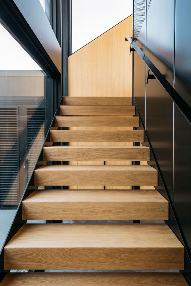 A close-up architectural photograph looking up a contemporary floating staircase. Broad warm timber treads are cantilevered from a dark structural frame, with slim black metal handrails, dark wall and window frames, glass balustrade, and light timber wall panelling. The stairwell is bright and vertically oriented, with strong geometric lines and a minimalist material palette.
