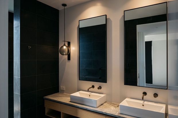 A contemporary double-vanity bathroom with two rectangular white vessel basins set on a long timber vanity with a dark stone benchtop. Large black-framed mirrors, dark charcoal wall tiles, warm neutral walls, black tapware, and a pendant light create a moody, refined interior.