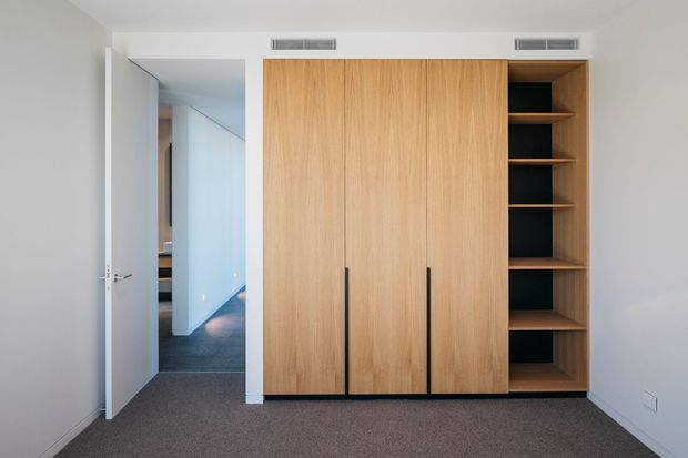 A minimalist built-in timber wardrobe and open shelving unit occupies one wall of a small, bright room. The joinery has pale wood sliding or hinged doors with long black vertical handles, dark open shelves, white plaster walls, grey carpet, and a partially open white door leading to a hallway.
