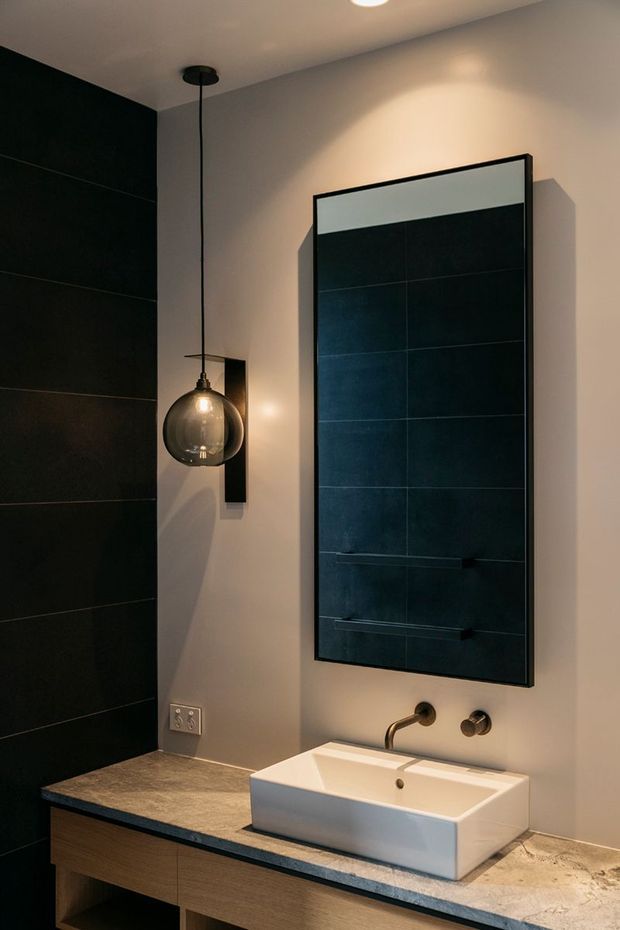 A contemporary bathroom vanity with a wall-mounted rectangular mirror, white vessel basin, dark stone-look benchtop, timber vanity cabinetry, large dark wall tiles, and a single globe pendant light. The warm neutral wall contrasts with the black tiled feature wall and bronze-toned tapware.