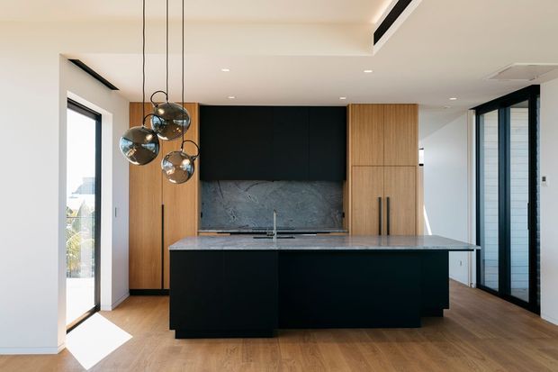 Contemporary kitchen featuring a large dark island, black integrated cabinetry, warm timber storage panels, a dark stone splashback, and pendant globe lighting. Full-height glazing and a timber-look floor create a bright, refined open-plan space.