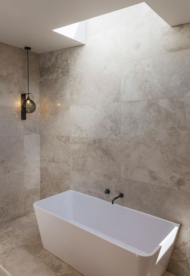 A contemporary bathroom featuring a freestanding white bathtub against large-format beige-grey stone-look wall tiles and matching floor tiles. A black wall-mounted bath tap and a suspended smoked-glass pendant light add contrast. A rectangular ceiling skylight brings in natural light.