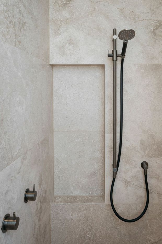 Close-up of a contemporary walk-in shower clad in large-format light beige stone-look tiles. A dark brushed-metal handheld shower set, hose, controls, and recessed tiled niche are visible.