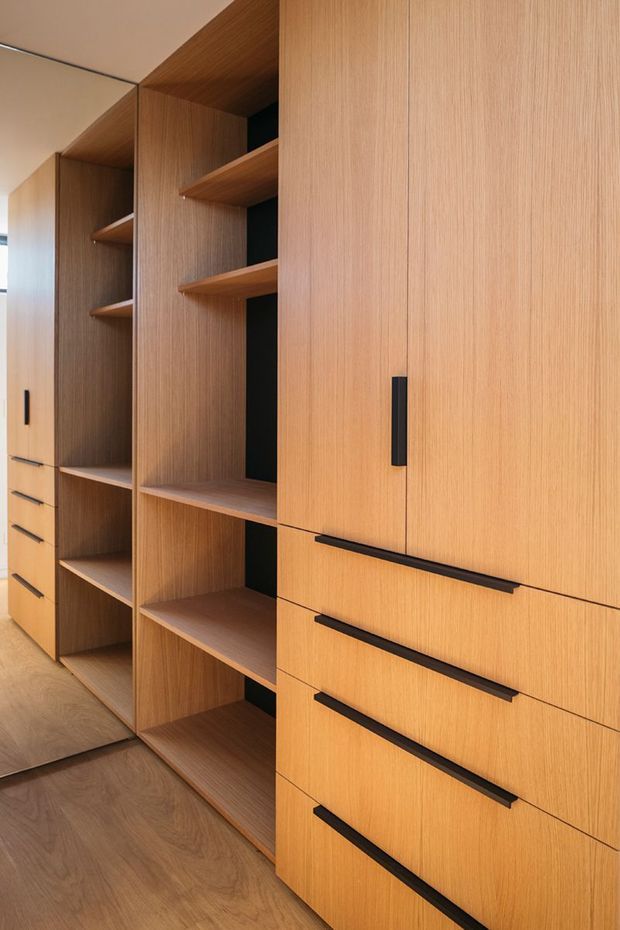 Close-up interior view of a wall-to-wall built-in storage system with warm wood-grain cabinetry, open shelving, tall cupboards, and multiple drawers fitted with slim black handles. A full-height mirror reflects the joinery, and the timber flooring continues through the space.
