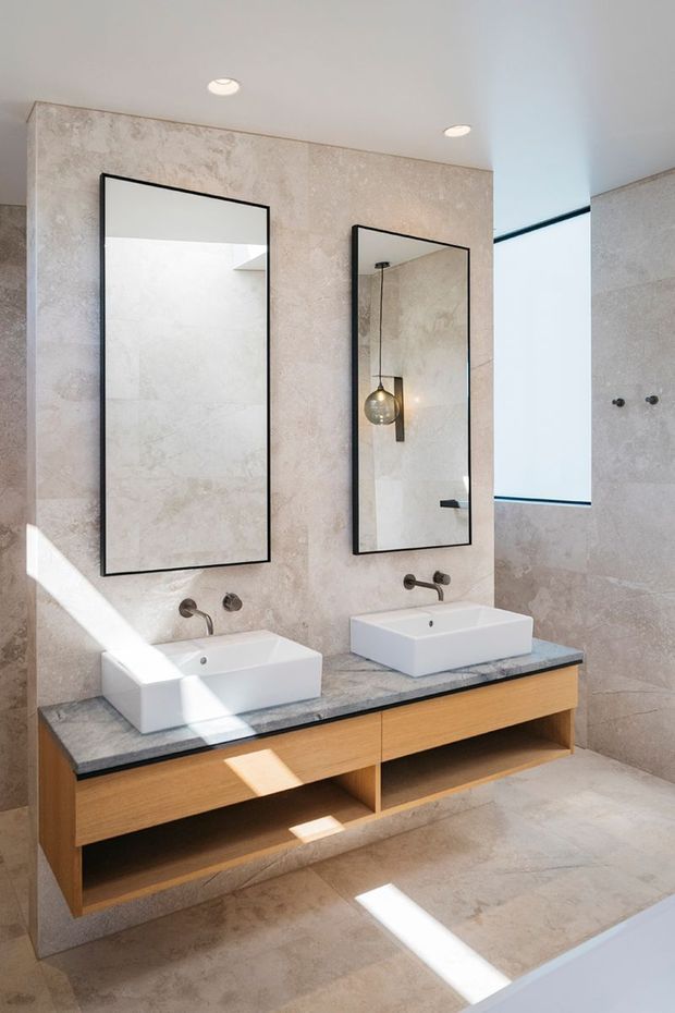 Contemporary bathroom with a floating double vanity, two white vessel basins, wall-mounted brushed metal taps, a dark stone benchtop, and warm timber drawers. Large pale stone-look wall and floor tiles create a clean, cohesive finish, while black-framed rectangular mirrors and a pendant light add contrast. Bright natural light enters from the right, with a partially visible shower area beyond.