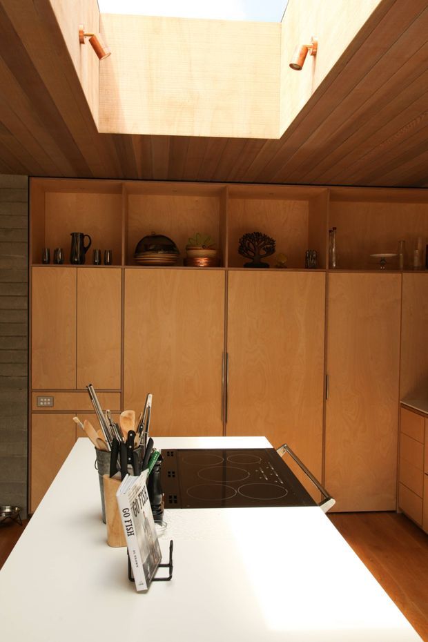 Warm timber-lined kitchen with extensive built-in plywood cabinetry, open display shelving, a white island benchtop and black induction cooktop. A large skylight illuminates the wood ceiling and cabinetry, while a dark timber-lined wall and cooking utensils reinforce the functional kitchen setting.