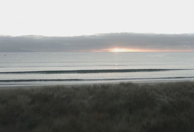 A subdued coastal landscape viewed across dense dune grass toward calm ocean water, with low layered waves and the sun emerging near the horizon beneath a heavy cloud bank. The image has a soft, muted, atmospheric quality and contains no visible building or interior space.