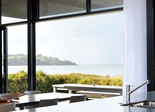 Bright contemporary kitchen and dining area opening onto a coastal outlook. The image is dominated by expansive floor-to-ceiling glazing with dark frames, a white kitchen island and stainless-steel mixer tap and sink in the foreground, plus a timber dining table and bench outside on the terrace. Ocean, shoreline vegetation and a distant headland are visible beyond the glazing.