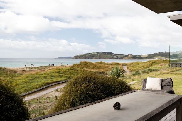 A coastal outdoor terrace or balcony overlooking a sandy beach, dune vegetation, calm sea, and a distant headland. The terrace has a large concrete or stone-topped table, cushioned outdoor seating, glass balustrading, and a projecting roof or ceiling above. A boardwalk leads through the dunes toward the beach.