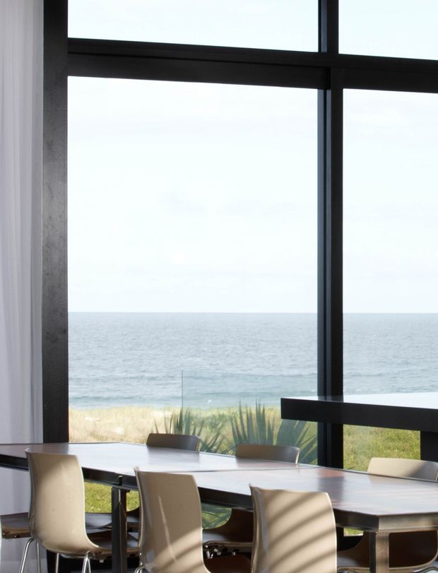 A contemporary dining area with a long rectangular table and upholstered dining chairs positioned beside expansive black-framed floor-to-ceiling glazing. The windows provide a direct view toward a sandy beachfront and ocean horizon, while sunlight creates strong horizontal shadows across the room. The space has a restrained, modern coastal feel with dark structural framing and pale furnishings.