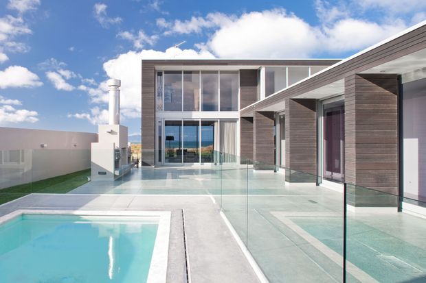 Modern multi-level residence viewed from an outdoor terrace, featuring a clear-glass pool barrier, swimming pool, pale tiled or concrete paving, extensive floor-to-ceiling glazing, and dark horizontal wood-look cladding. The flat-roofed composition includes a covered outdoor corridor and a bright blue-sky setting.