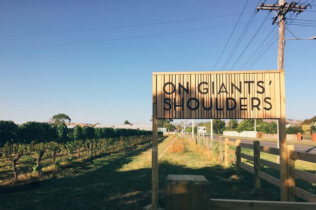 A rural roadside scene featuring a large timber-framed sign reading “ON GIANTS’ SHOULDERS,” set beside a vineyard or agricultural rows. A timber fence, utility poles, open fields, and distant low hills are visible under a clear blue sky.