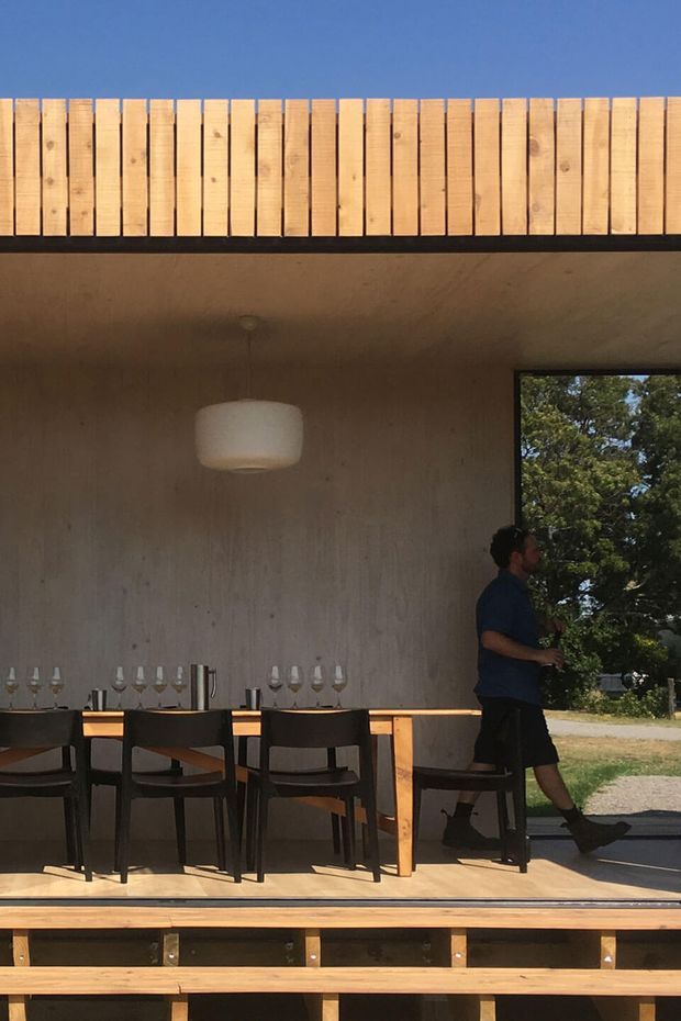 A covered outdoor dining and entertaining area with a long timber dining table, dark framed chairs, wine glasses, a pendant light, and a compact bar or serving bench. The space has raw concrete walls and ceiling, extensive timber decking and a timber slatted parapet or privacy screen. A large opening frames trees and the surrounding landscape, creating a relaxed indoor-outdoor setting.