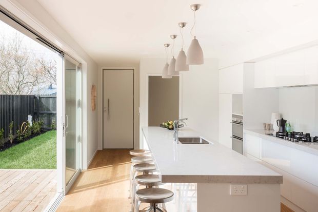 Bright, minimalist contemporary kitchen viewed from an open living area, featuring a long white island with integrated sink and stools, white full-height cabinetry, pendant lights, timber flooring, and large sliding glass doors opening to a small landscaped outdoor area.
